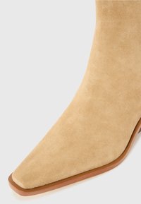 Stivale ankle in suede beige con punta affilata e suola sottile marrone. Texture liscia senza hardware o motivi visibili.