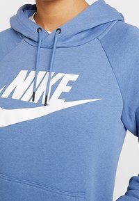 Blå hoodie i mjukt tyg med en stor vit Nike-logotyp framtill, snoddar med svarta spetsar och en känguruficka.