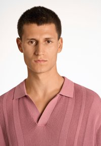 Růžový polo tričko s V-výstřihem a žebrovanou texturou, s krátkým rukávem a uvolněným střihem. Jednoduchý design bez vzorů nebo akcentů.