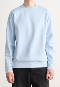 Sweatshirt bleu clair à manches longues, avec un col côtelé et des poignets. Présente un petit logo sur la poitrine. Tissu doux avec une texture lisse.