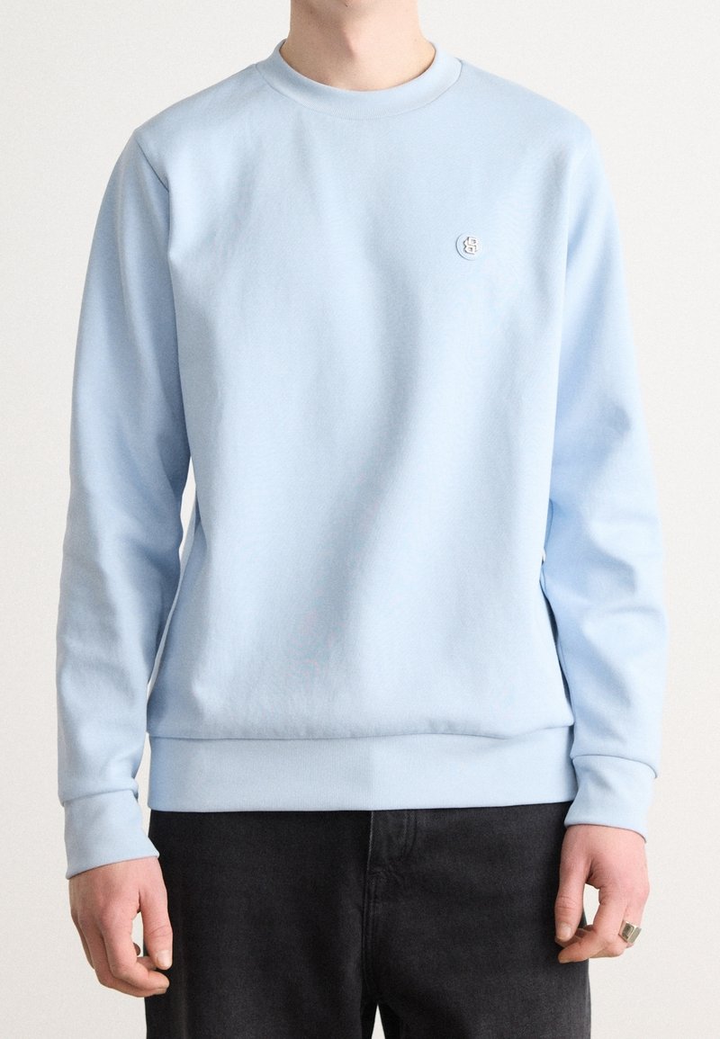 Sweatshirt bleu clair à manches longues, avec un col côtelé et des poignets. Présente un petit logo sur la poitrine. Tissu doux avec une texture lisse.