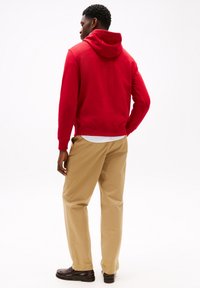 Red hooded sweatshirt with a relaxed fit, paired with beige trousers and dark shoes. Features ribbed cuffs and hem.
