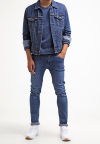 Giubbotto di jeans blu con polsini piegati, felpa blu navy sotto, jeans blu e scarpe da ginnastica bianche con dettagli beige. Completo casual.