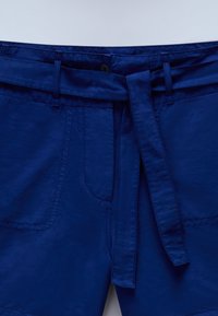 Pantaloni corti in cotone blu con vita regolabile, due tasche frontali e dettagli di cucitura visibili. Il tessuto ha una texture liscia.