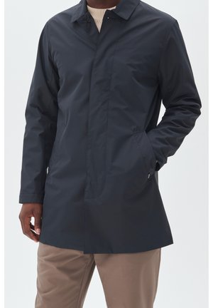 Mies pukee tummansinisen sadeviitan beigen neuleen ja vaaleanruskeiden housujen päälle, toinen käsi taskussa, yksinkertaista vaaleaa taustaa vasten.