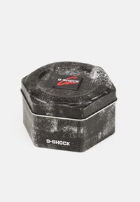 Boîte en métal noire hexagonale avec une surface texturée, arborant un logo G-SHOCK rouge et blanc sur le dessus et de simples accents argentés autour du bord.