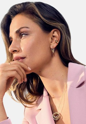 Guld hoopsøreringe, lagdelt cirkelhalskæde, blød pink blazer, bølget brunt hår, glat hud, naturlig makeup, neutral baggrund.