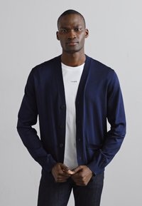 Cárdigan azul marino hecho de un material de punto suave, con cierre de botones en la parte frontal, dos bolsillos laterales y un corte relajado, usado sobre una camiseta blanca.