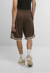 Shorts de sport bruns avec des rayures créme contrastantes au niveau de l'ourlet, fabriqués en tissu lisse, avec une taille élastique. Portés avec des chaussettes blanches et des baskets.