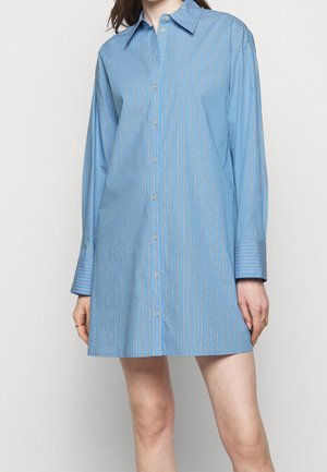 Kobieta ubrana w niebieską sukienkę-koszulę z długim rękawem, zapinaną na guziki, z cienkimi pionowymi białymi i różowymi paskami.