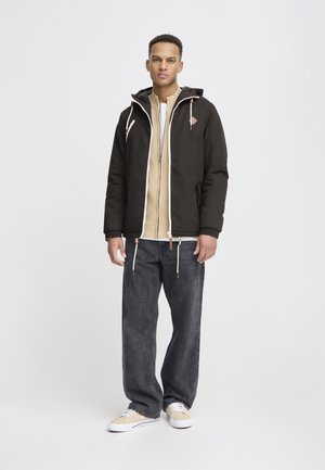 Schwarze Kapuzenjacke mit Kordelzügen, beigem Innenfutter, grauen Jeans und hellen Sneakers. Die Jacke hat ein Logo und Reißverschlüsse.
