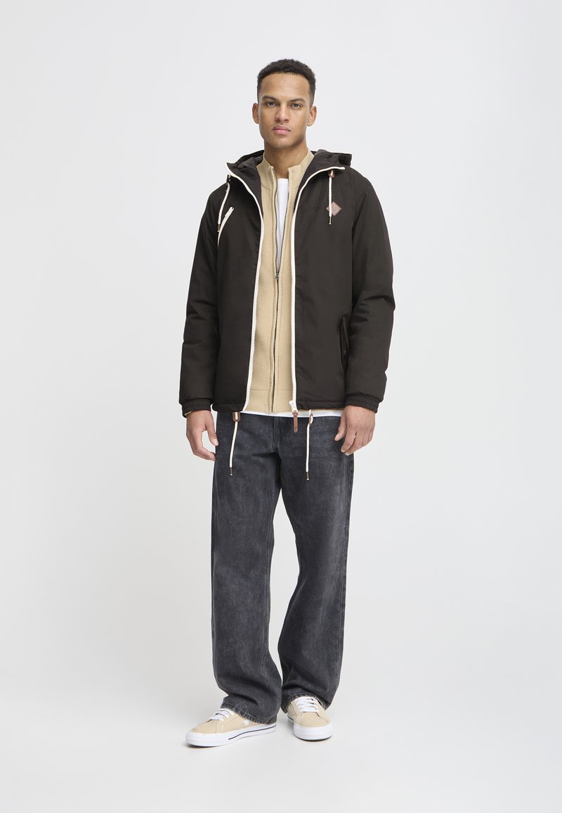 Black hooded jacket with drawstrings, beige inner layer, gray jeans, and light-colored sneakers. The jacket features a logo and zippers.
