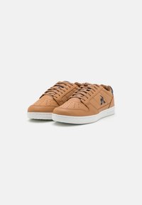 Sneaker in pelle marrone chiaro con suole in gomma bianca, caratterizzate da lacci rotondi, dettagli perforati sulla punta e un accento blu navy nel tallone.