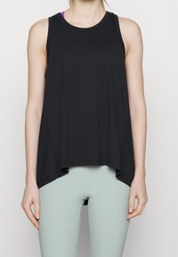 Žena oblečená do bezrukávového černého asymetrického sportovního topu a světle zelených přiléhavých legín, stojící na jednobarevném pozadí.