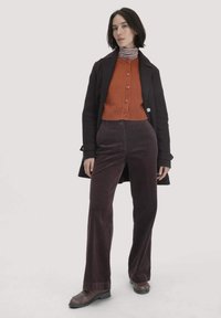 Vrouw in een donkere jas, roestoranje knoopcardigan over een gestreept turtleneck, donkere corduroy broek en donkerbruine loafers, staand tegen een witte achtergrond.