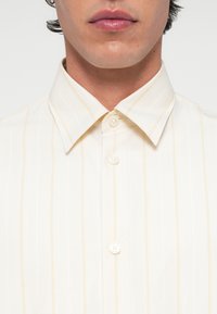 Férfi krém színű ingben, amelyen finom, függőleges sárga csíkok vannak, és három gomb látható elöl.