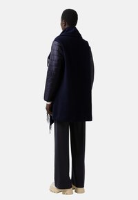 Námořnický modrý kabát s prošívanými tmavě fialovými rukávy, s třásněmi na lemu, v kombinaci s širokými kalhotami a béžovými botami s masivní podešví.