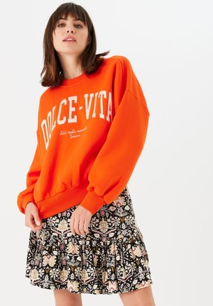Oranje sweatshirt met "DOLCE-VITA" in het wit gedrukt, gecombineerd met een zwarte rok met een bloemmotief en ruffles, in verschillende kleuren en vormen.