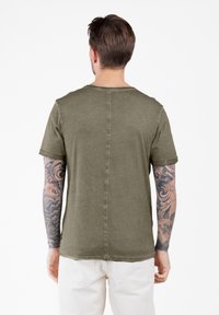 Camiseta de algodón verde oliva con mangas cortas, dobladillo sin terminar y un corte holgado, que presenta una costura central y puños de manga enrollados.