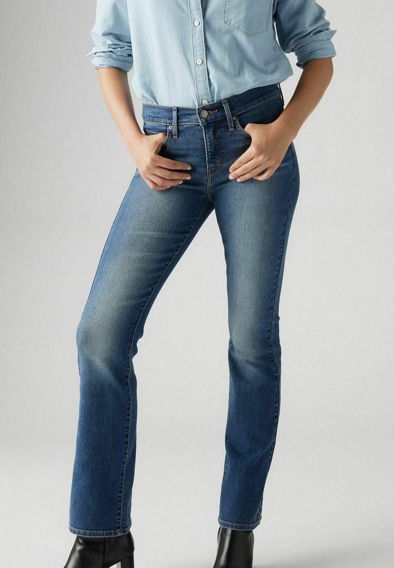 Dunkelblaue Jeans im Bootcut-Design, mit einer mittelhohen Taille, fünf Taschen und leichtem Ausbleichen an den Oberschenkeln. Getragen mit einem hellblauen Hemd.