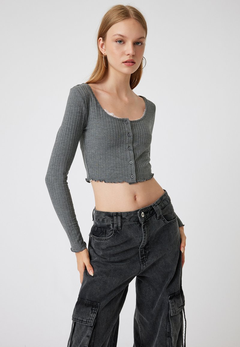 Top corto de manga larga gris con relieve, escote amplio y detalle de encaje, que presenta un botón y un dobladillo sin terminar, combinado con pantalones cargo negros.