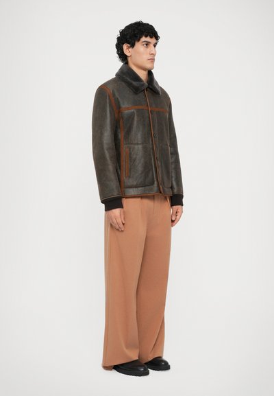 Bruine leren jas met een bontkraag, voorzien van oranje stiksels en knopen, gecombineerd met losse, lichtbruine broek en zwarte schoenen.