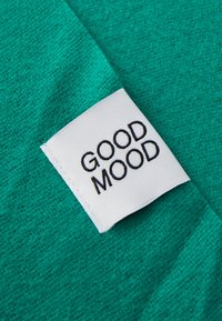 Tessuto turchese con una trama testurizzata e un'etichetta bianca con la scritta "GOOD MOOD" in caratteri neri, cucita nell'angolo.