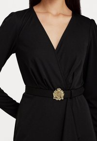Svart omslagklänning med djup V-ringning, lång ärm och en texturerad guldspänne med ett emblem. Slät tyg med en figursydd design.