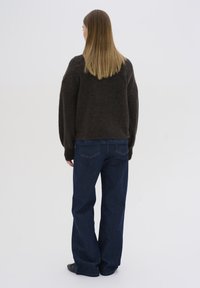 Braun gestrickter Pullover mit fallenden Schultern, kombiniert mit dunkelblauen Schlagjeans. Modell von hinten fotografiert, zeigt die Rückendetails beider Kleidungsstücke.