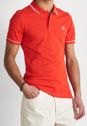 Mann trägt rotes Poloshirt mit kurzen Ärmeln und weißem Besatz sowie weiße Hose, eine Hand in der Tasche und braunes Armband am linken Handgelenk.