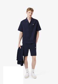 Set di camicia a maniche corte e pantaloni corti di colore blu navy realizzato in tessuto testurizzato, con un piccolo logo verde. Il modello tiene anche una giacca abbinata.