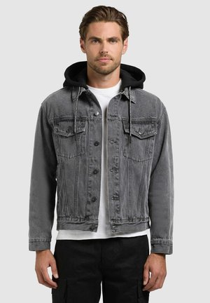 Mand iført grå denimjakke med sort hætte, hvid t-shirt og sorte bukser, stående foran en ensfarvet lys baggrund.