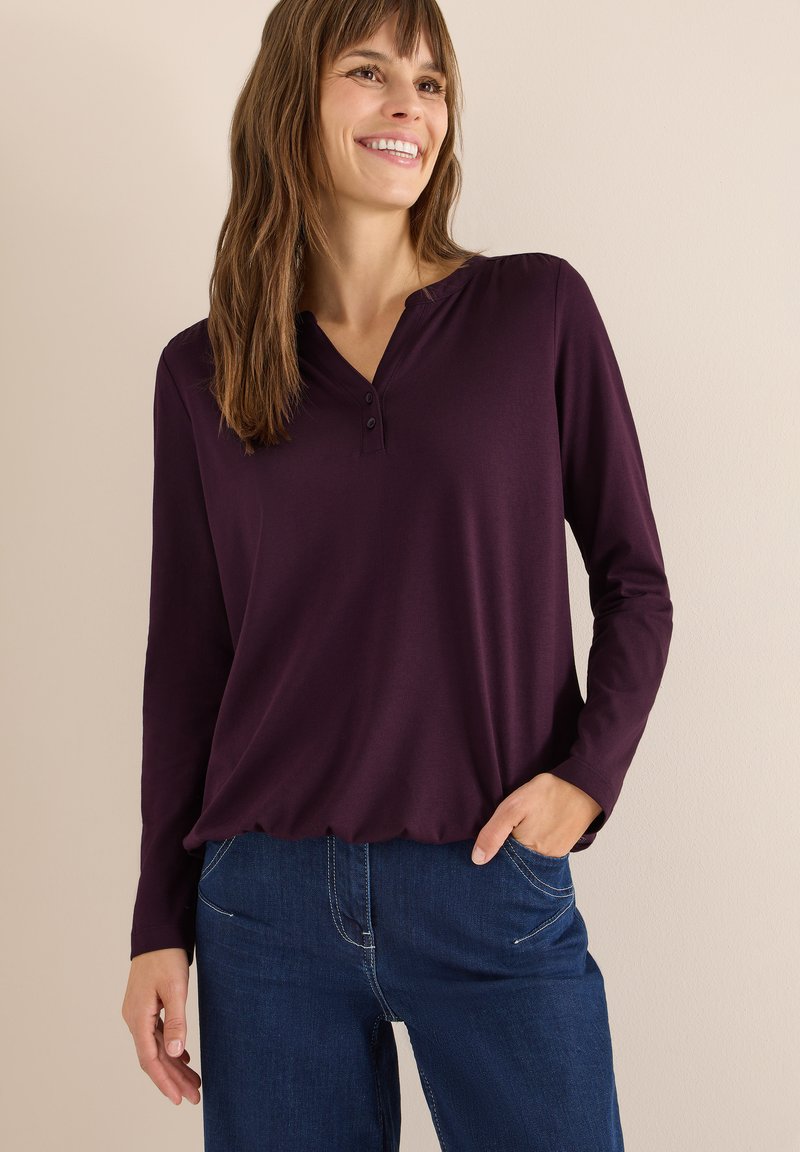 Langsleeve henley-stijl top in diep paars, met een ronde hals, drie knopen en een relaxte pasvorm. Gecombineerd met donkerblauwe jeans.