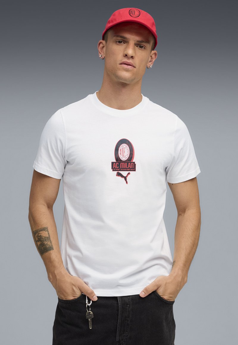Weißes Baumwoll-T-Shirt mit AC Milan-Logo und roten Puma-Akzenten, kombiniert mit einer passenden roten Mütze, getragen mit schwarzen Hosen und Schlüsseln.