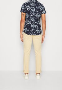 Camicia a maniche corte blu navy con motivo floreale bianco, abbinata a chinos beige chiaro e sneakers bianche, vista da dietro.