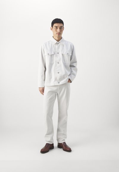 Veste en denim blanche avec deux poches poitrine, associée à un pantalon blanc et des bottines marron. Le mannequin se tient devant un fond uni.