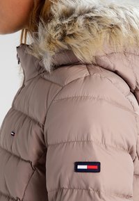Vadderad beige jacka med huva i fuskpäls, med horisontell sömnad, en logotyp patch på ärmen och dragkedjestängning.