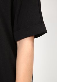 Musta lyhythihainen paita, jossa on käännetyt hihansuut; valmistettu sileästä, teksturoidusta kankaasta. Näyttää osan käsivarresta neutraalia taustaa vasten.