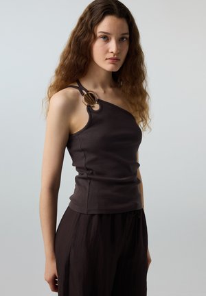 Giovane donna con capelli lunghi e mossi che indossa un top marrone scuro senza maniche con un anello decorativo su una spalla, abbinato a pantaloni scuri.