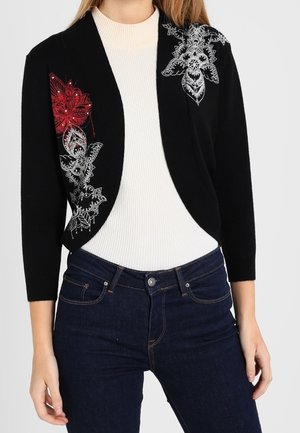 Schwarzer kurzer Cardigan mit roter und weißer Blumenstickerei, getragen über einem weißen, gerippten Oberteil und dunkelblauen Jeans.