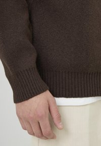 Pull en tricot marron avec des poignets côtelés, un tissu texturé et une coupe décontractée. Superposé sur une chemise blanche et associé à un pantalon en velours côtelé clair.