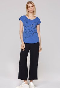 Blauwe katoenen t-shirt met ronde hals en korte mouwen, voorzien van zwarte cursieve tekst. Gecombineerd met wijde zwarte broek en witte sneakers.
