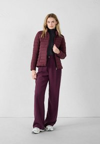 Bordeauxkleurige pufferjack met een gequilt textuur, ritsluiting en hoge kraag, gecombineerd met losse donkerpaarse broeken en grijze sneakers.