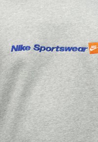 Γκρί ύφασμα με μπλε κείμενο "Nike Sportswear" και πορτοκαλί τετράγωνο λογότυπο Nike με λευκή σβαστικοειδή γραμμή στα δεξιά.