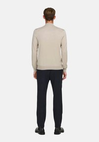 Suéter beige claro con mangas largas, puños y cuello acanalados; combinado con pantalones oscuros y zapatos negros, visto desde la parte posterior.