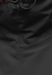 Černá látka bundy s texturovaným povrchem, která má stahovací šňůrku na krku a viditelné švy.