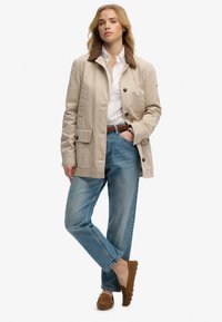 Mujer con chaqueta beige de cuello oscuro, camisa blanca abotonada, vaqueros azules, cinturón marrón y mocasines marrones, de pie con una mano en el bolsillo.