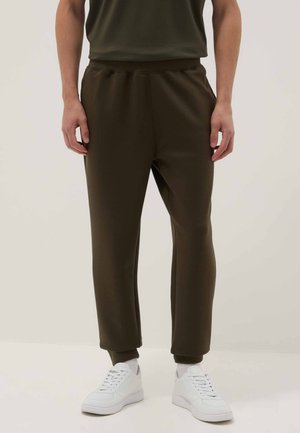 Personne portant un pantalon de jogging marron foncé et des baskets blanches, debout devant un fond clair uni.