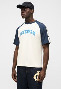 Ανδρικό κοντομάνικο μπλουζάκι σε κρεμ χρώμα με ναυτικό μπλε λεπτομέρειες, με το λογότυπο "ICECREAM" σε μπλε γράμματα και σχεδιασμό λογότυπου στα μανίκια.