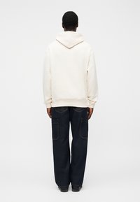 Μπεζ φουτέρ με μεγάλο καπό στην πλάτη, σε συνδυασμό με άνετο σκούρο τζιν παντελόνι cargo με πολλές τσέπες και έντονη αντίθεση στα ραφές.